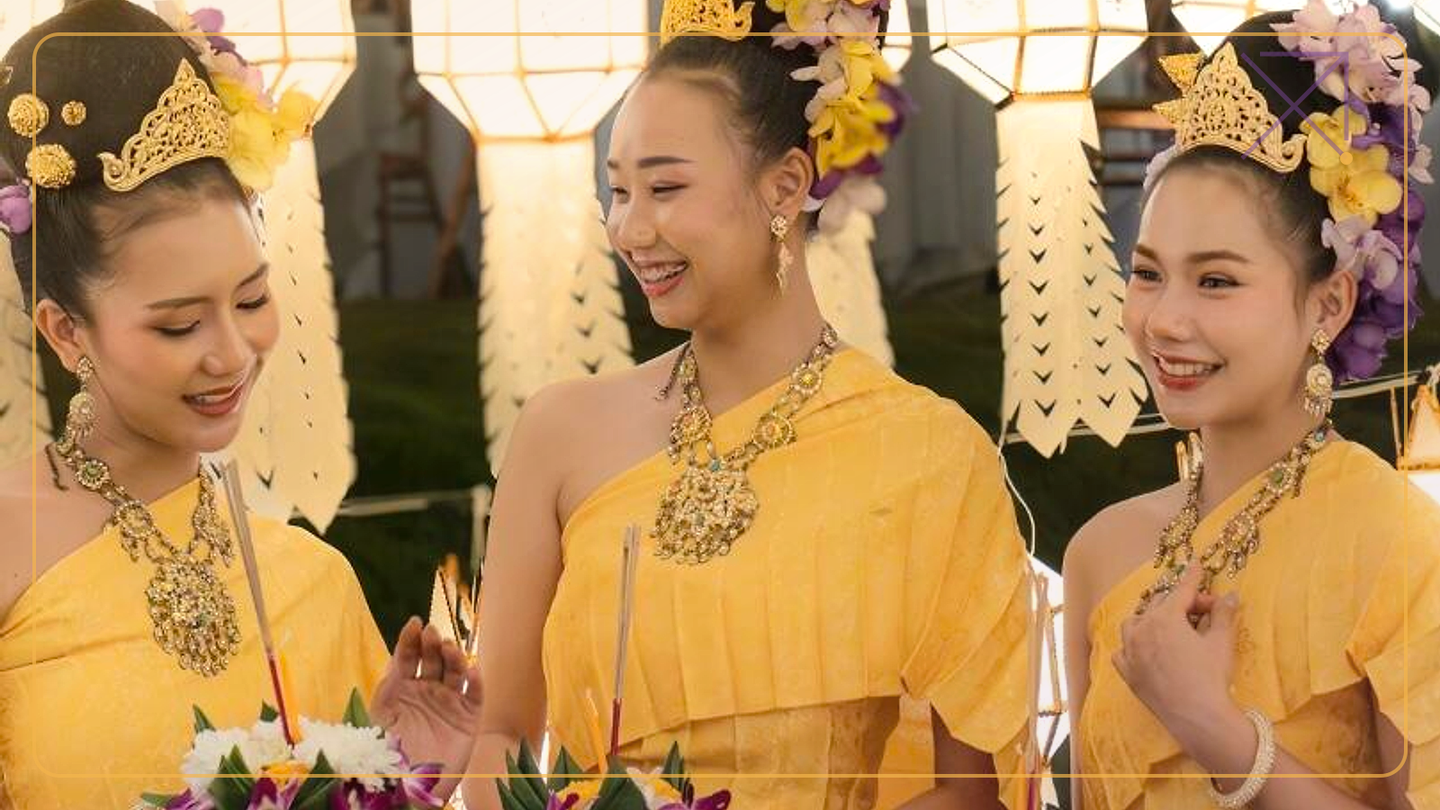 Three women in traditional dress hold decorated krathongs beneath hanging lanterns